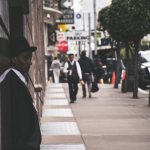 A man in a suit and hat leans against a city building, observing pedestrians. — The Hidden Mystery Behind B2B Marketing Agenc